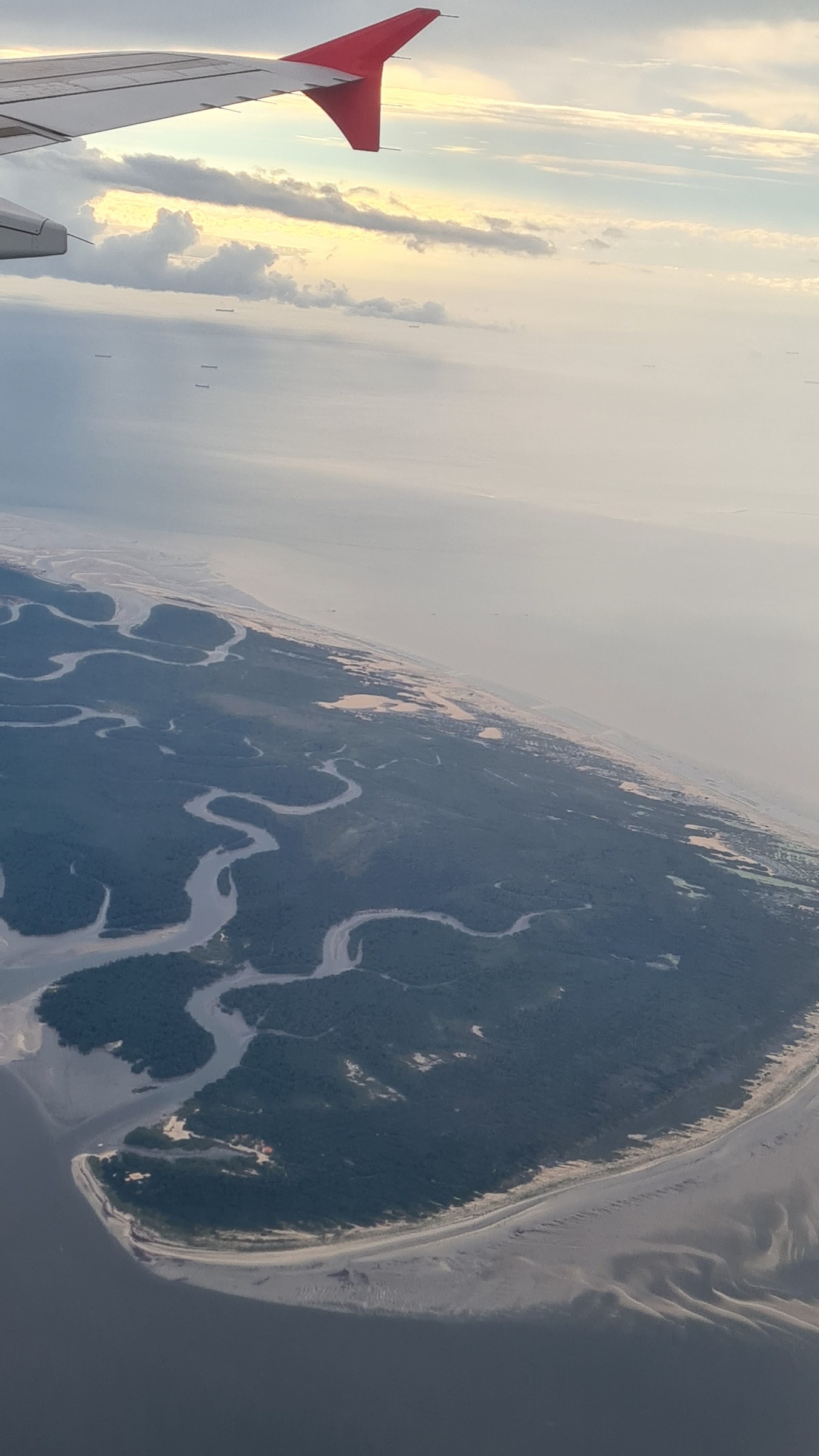 Aerial view from an airplane window at sunset over the Parnaíba Delta, dozens of mangrove islands cut by winding tidal channels meeting the Atlantic Ocean, red winglet in frame