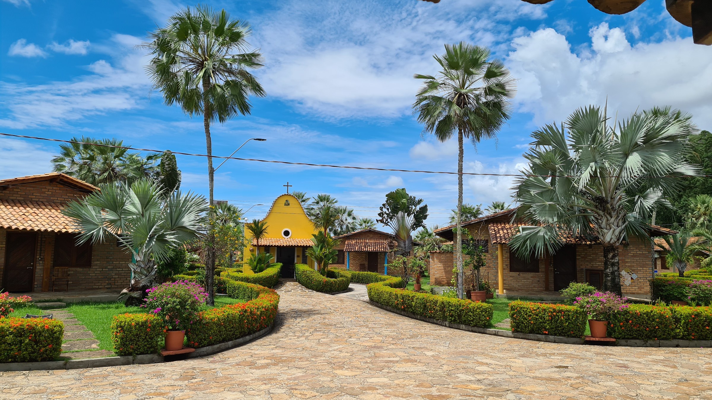 Stone walkway lined with palm trees and flowering hedges leading to a small yellow chapel at Porto Preguiças Resort, red-brick tile-roofed bungalows on either side