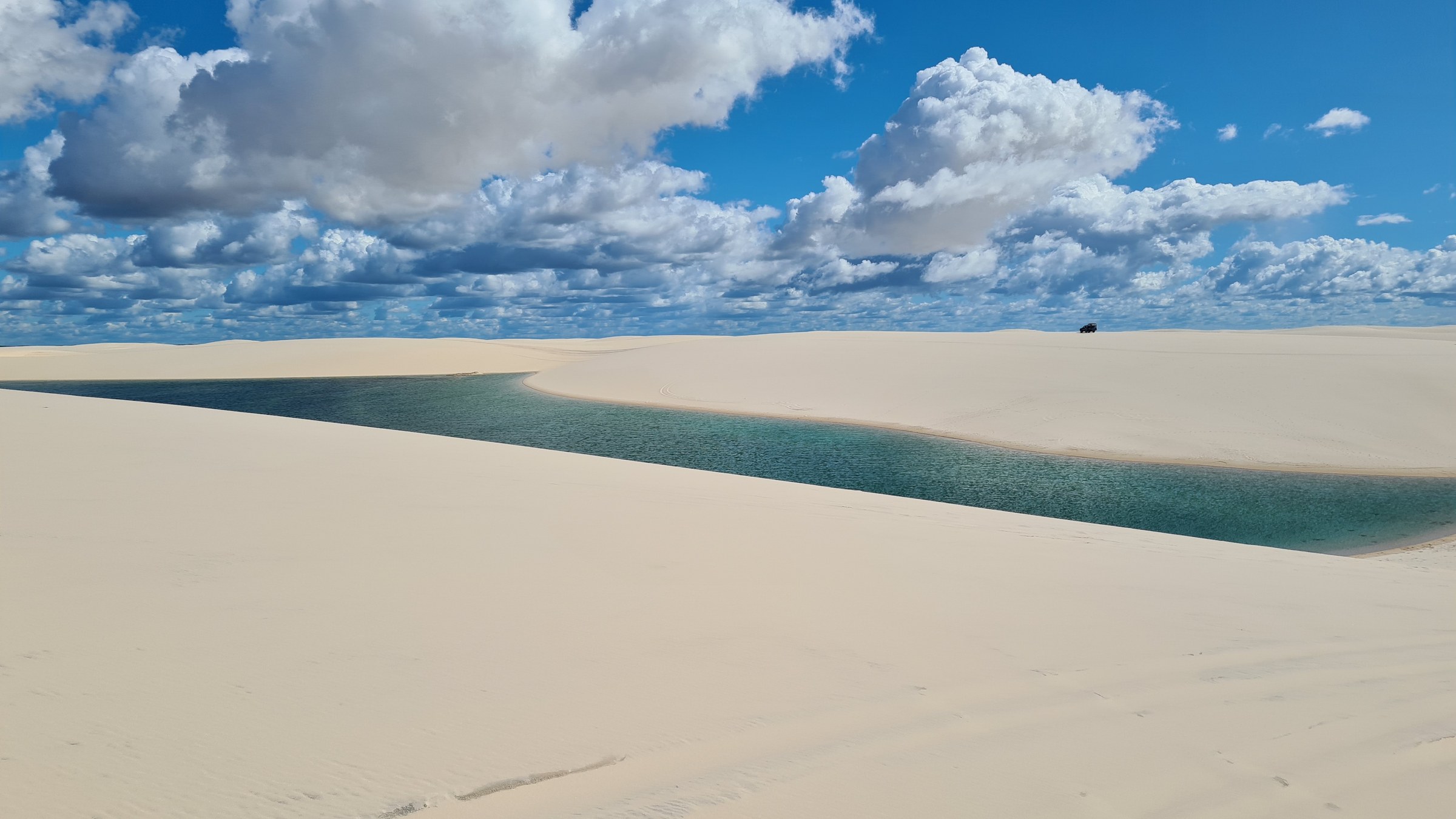 Turquoise lagoon winding between dunes with a dune buggy visible as a small silhouette on the horizon