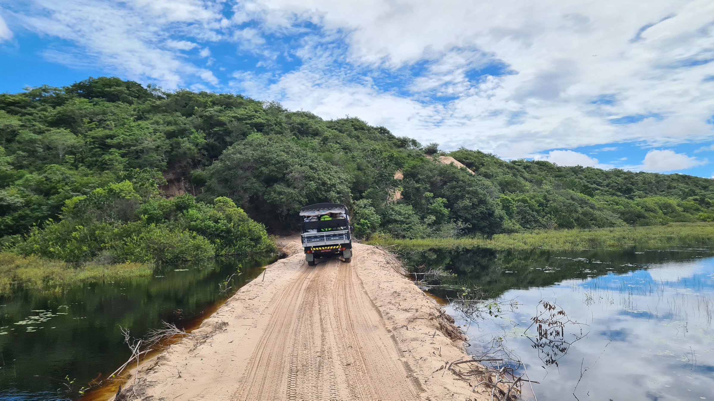 A 4x4 buggy crossing a narrow sandy causeway between two flooded pools, forested hills ahead, near Ponta do Mangue
