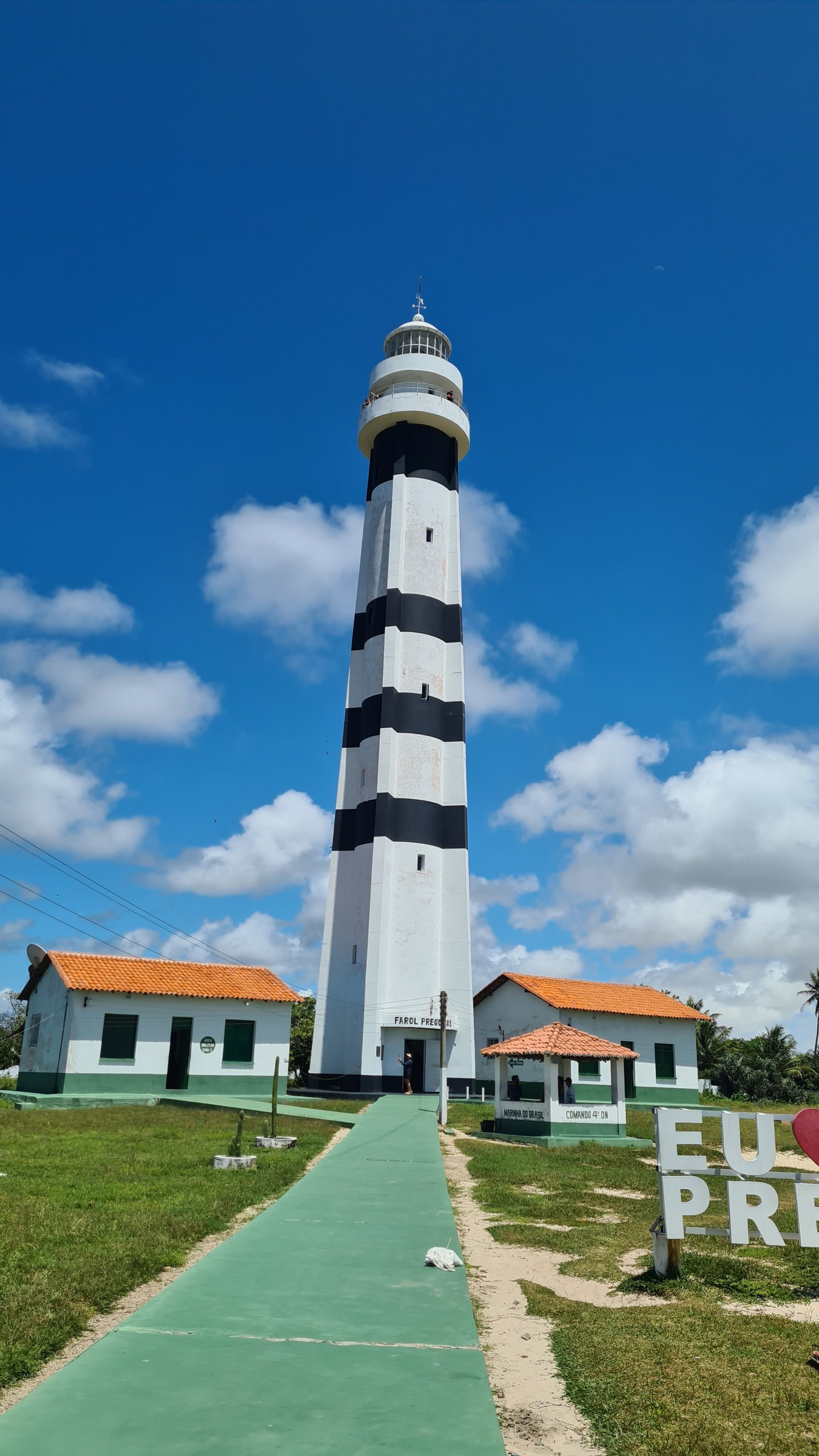 Black and white striped Farol de Mandacaru lighthouse seen head-on from base to top against a blue sky, small green-and-white outbuildings at its base, palm-lined walkway leading up to it