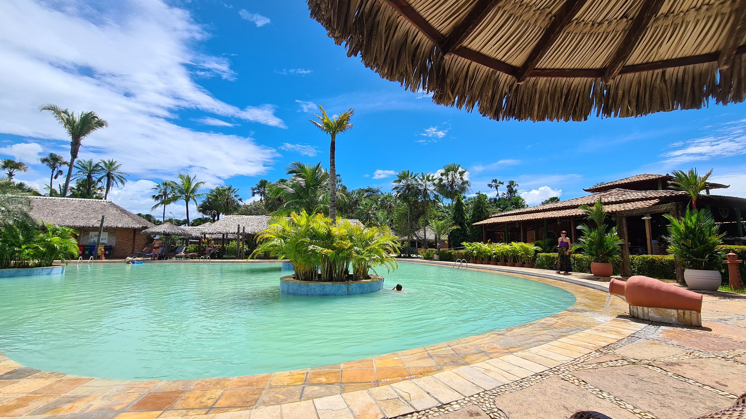 Turquoise pool at Porto Preguiças Resort under a thatched umbrella, a palm-planted island at its center, thatched-roof buildings and palm trees around the deck