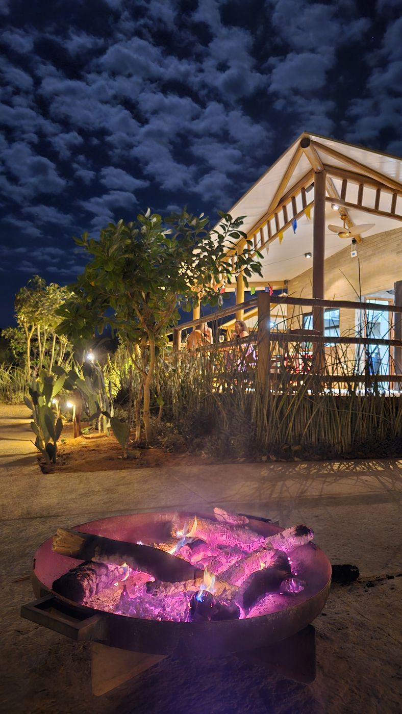Fire pit glowing at night under a dramatic cloudy sky, main house behind, Sertão do Luar